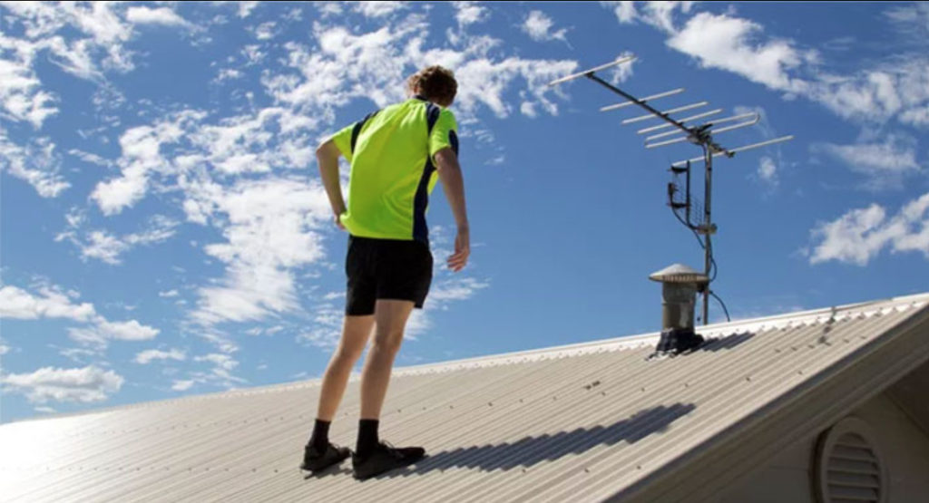 A man in a yellow shirt at the top of the roof, completing a roof inspection